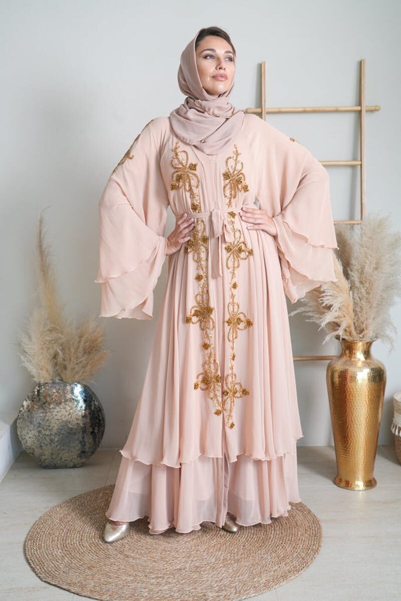 Photo by Qalanjos Fashions -Online Abaya Store Studio portrait of a woman wearing a pink abaya with golden embroidery and a headscarf.
